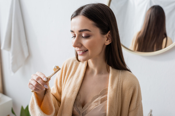 young and cheerful woman in bathrobe looking at toothbrush with toothpaste  - Fotó, kép