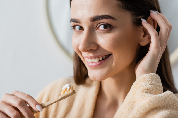 portrait of cheerful woman in bathrobe holding toothbrush with toothpaste while adjusting hair  - Foto, afbeelding