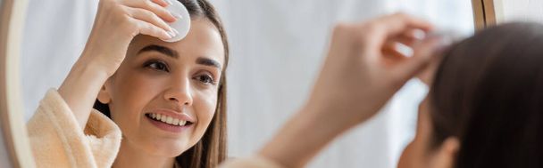 αντανάκλαση χαμογελαστή νεαρή γυναίκα καθαρισμού πρόσωπο με βαμβάκι μαξιλάρι στο μπάνιο καθρέφτη, πανό  - Φωτογραφία, εικόνα
