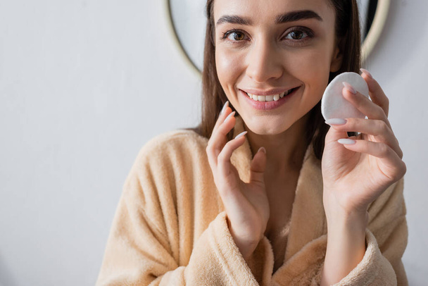 cheerful young woman in bathrobe holding clean cotton pad in bathroom  - Fotografie, Obrázek
