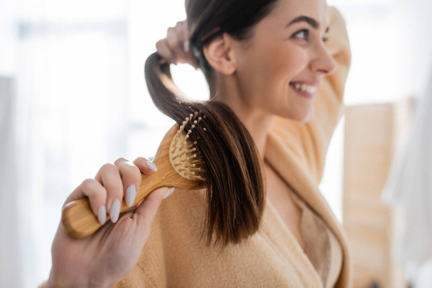 sorrindo e borrada jovem mulher escovando cabelo brilhante no banheiro  - Foto, Imagem