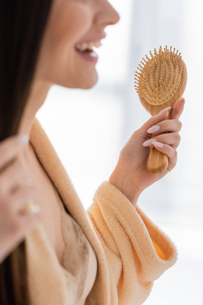 vista ritagliata di felice giovane donna in possesso di spazzola per capelli in legno in bagno  - Foto, immagini