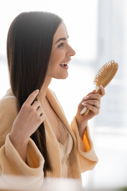side view of happy young woman holding wooden hair brush in bathroom  - Foto, Bild