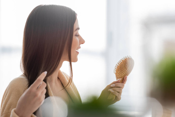 side view of cheerful young woman holding wooden hair brush in bathroom  - Foto, Imagen