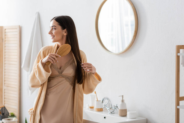 cheerful young woman in bathrobe brushing hair in white modern bathroom  - Foto, immagini
