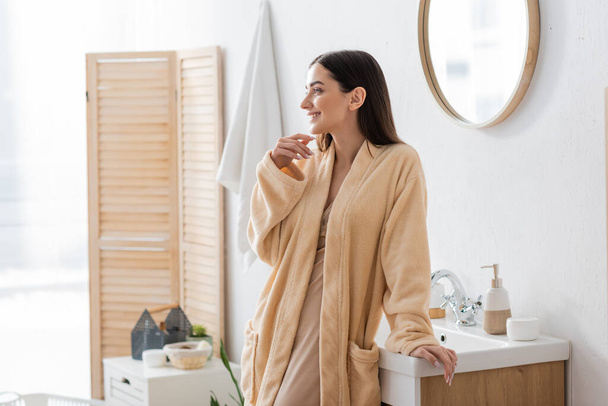 happy young woman in bathrobe looking away in modern bathroom  - Foto, afbeelding
