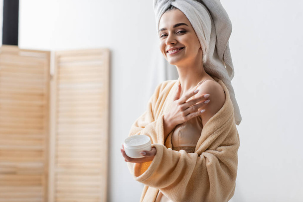 mujer joven complacida con toalla en el recipiente de retención de la cabeza mientras aplica mantequilla corporal  - Foto, Imagen