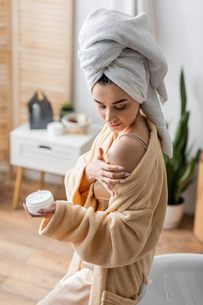 young woman in bathrobe with towel on head holding container while applying body butter  - 写真・画像