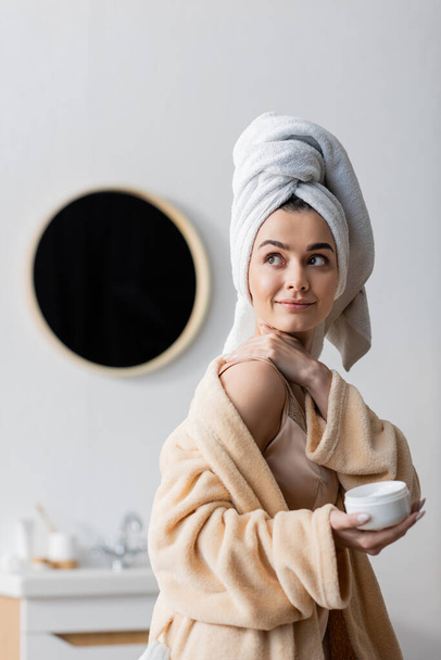 Smiling woman in towel and bathrobe applying cosmetic cream in bathroom  - Фото, зображення