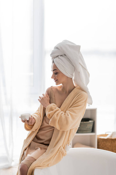 Вид сбоку улыбающейся женщины в полотенце и халате, наносящей косметический крем возле ванной дома  - Фото, изображение