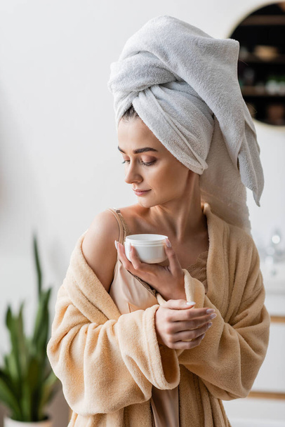 Young woman in towel on head and bathrobe holding cosmetic cream  - Foto, immagini