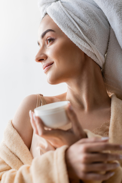 Portrait of pleased woman in towel and bathrobe holding blurred cosmetic cream in morning  - Foto, Bild