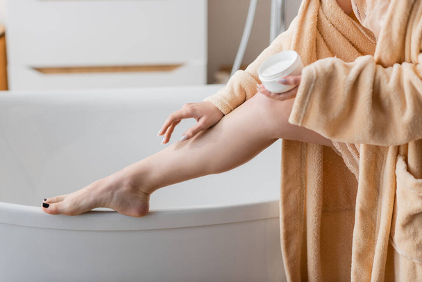 Cropped view of young woman applying cosmetic cream on leg near bathtub  - Photo, image