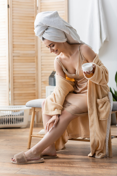 Vrolijke jonge vrouw in badjas en slippers aanbrengen crème op been in de badkamer  - Foto, afbeelding
