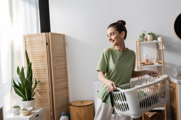 Fröhliche junge Frau hält Korb mit Kleidung in Waschküche  - Foto, Bild