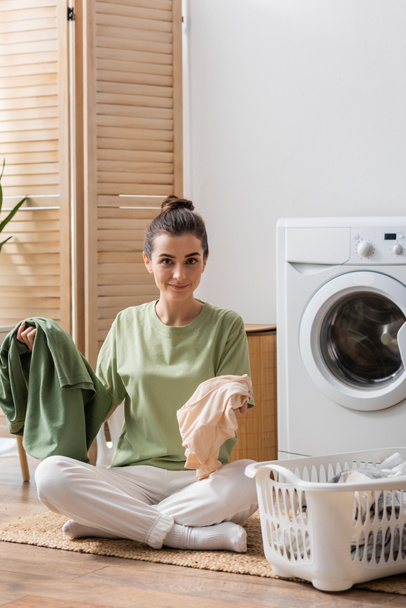Θετική γυναίκα κρατώντας ρούχα κοντά στο καλάθι και πλυντήριο στο σπίτι  - Φωτογραφία, εικόνα