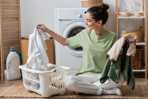 Щаслива молода жінка сортує одяг біля пральної машинки в пральні
  - Фото, зображення
