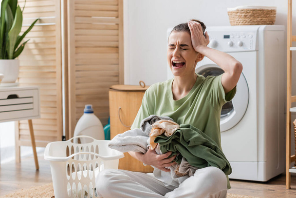 αναστατωμένη γυναίκα κρατώντας ρούχα κοντά στο καλάθι και κλαίγοντας στο πλυσταριό  - Φωτογραφία, εικόνα