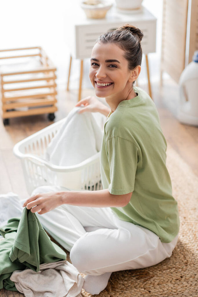 Cheerful young woman looking at camera near clothes and basket on floor in laundry room  - Fotografie, Obrázek