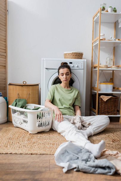 Tired woman sitting near clothes and washing machine in laundry room  - Foto, Bild