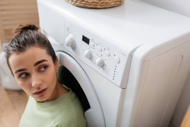 Брюнетка смотрит на стиральную машину в прачечной  - Фото, изображение