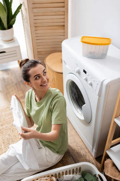 High angle view of smiling woman looking at box on washing machine near basket with clothes in laundry room  - Фото, зображення