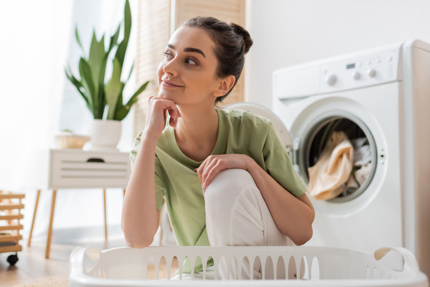Verträumte Frau sitzt neben Korb und verschwommener Waschmaschine zu Hause  - Foto, Bild