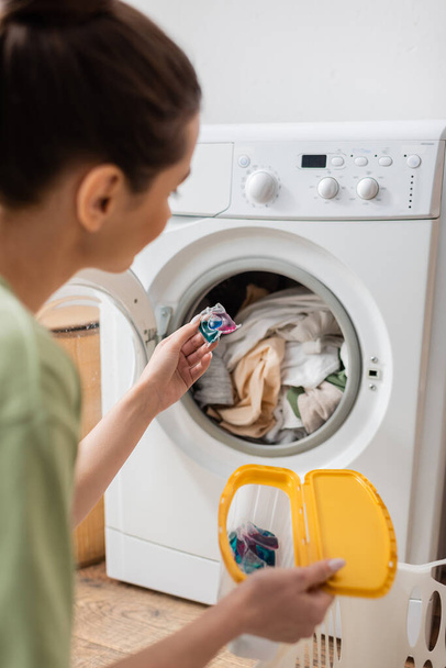 Blurred young woman holding washing capsule near machine in laundry room  - Foto, afbeelding