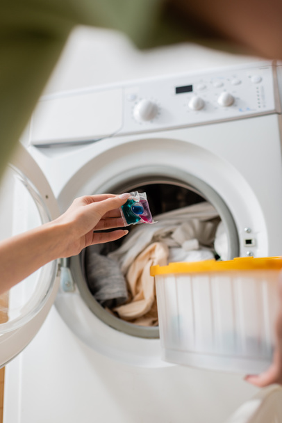 Cropped view of woman holding detergent pod near washing machine with laundry  - Foto, afbeelding