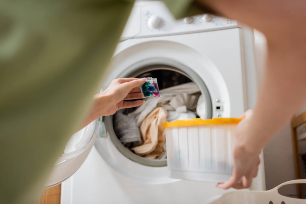 Cropped view of woman holding liquid detergent pod near clothes in washing machine at home  - Foto, afbeelding