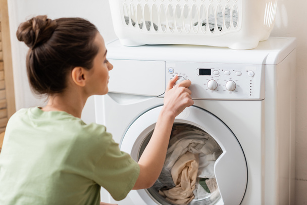Side view of blurred woman switching washing machine near basket in laundry room  - 写真・画像