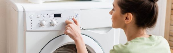 Вид сбоку на молодую женщину, переключающую стиральную машину дома, баннер  - Фото, изображение