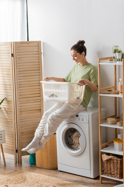 Young woman looking at basket while sitting on washing machine in laundry room  - Foto, afbeelding