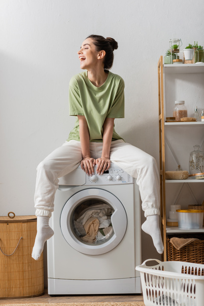 Возбужденная молодая женщина сидит на стиральной машине в прачечной  - Фото, изображение