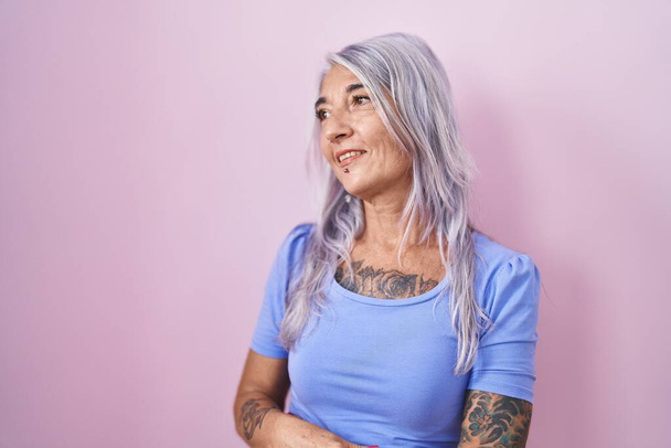 Жінка середнього віку з татуюваннями, що стоять над рожевим тлом, дивиться в сторону з посмішкою на обличчі, природним виразом. впевнений сміх
.  - Фото, зображення