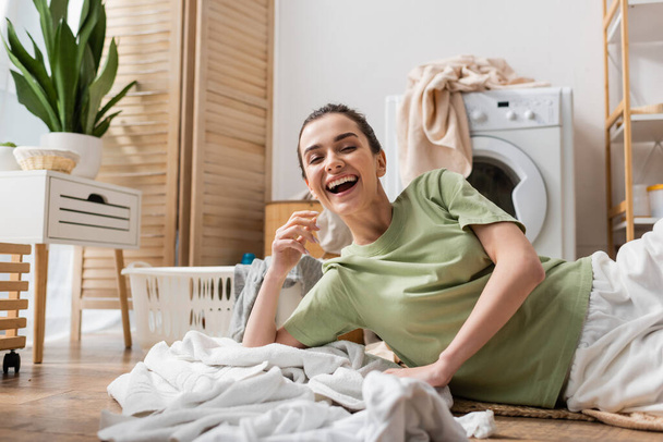 Fröhliche Frau liegt neben Kleidung und Korb in Waschküche auf dem Boden  - Foto, Bild