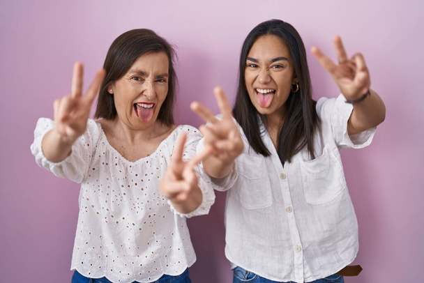 Hispanic mother and daughter together smiling with tongue out showing fingers of both hands doing victory sign. number two.  - Zdjęcie, obraz