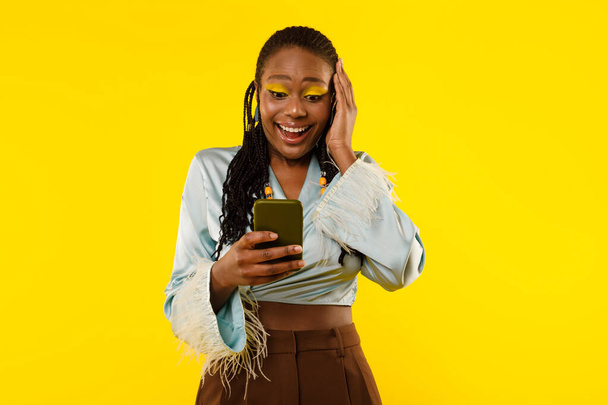 Excited African American Woman Using Smartphone Texting And Reading Wow Mobile Offer Standing On Yellow Studio Background. Great Mobile Application Advertisement Banner - Foto, Bild