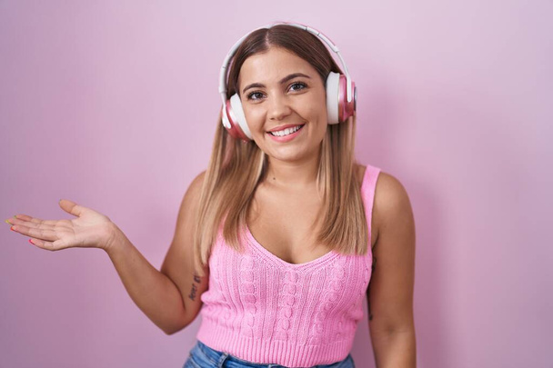 Young blonde woman listening to music using headphones smiling cheerful presenting and pointing with palm of hand looking at the camera.  - Foto, Imagem