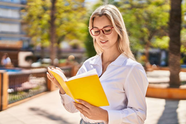 jonge blonde vrouw glimlachen zelfverzekerd lezen boek in park - Foto, afbeelding