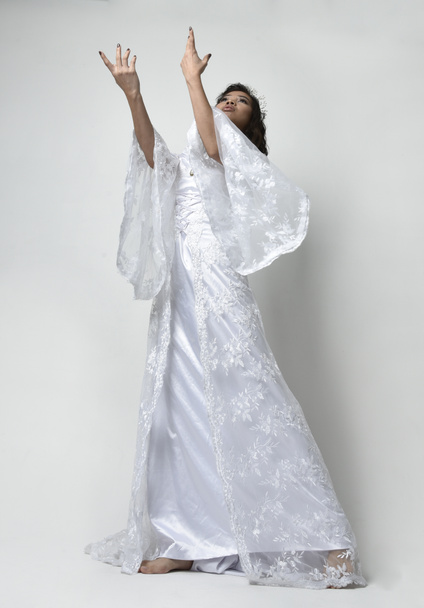 Full length portrait of beautiful woman wearing  fantasy costume, white bridal gown.  Standing pose with low angle perspective,  gestural arm movements casting a spell. Isolated white studio background - 写真・画像