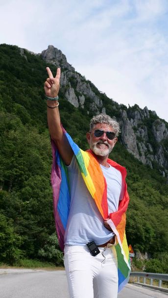 седовласый пожилой кавказский мужчина бисексуальности, диабетик с бородой и солнцезащитные очки с радужным ЛГБТКИА мирным флагом в горах. Празднует месяц гордости, День радужного флага, гей-парад - Фото, изображение