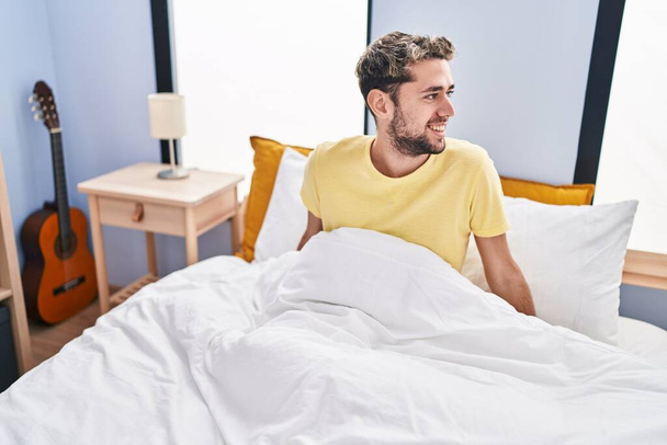 Νεαρός άνδρας χαμογελά σίγουρος κάθεται στο κρεβάτι στο υπνοδωμάτιο - Φωτογραφία, εικόνα