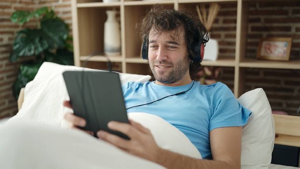 Young hispanic man using touchpad and headphones lying on bed at bedroom - Photo, image