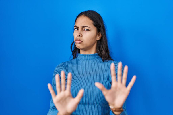Młoda brazylijska kobieta stojąca nad niebieskim, odizolowanym tłem odsuwając dłonie, pokazując odmowę i zaprzeczenie ze strasznym i obrzydliwym wyrazem twarzy. zatrzymać i zabronione.  - Zdjęcie, obraz