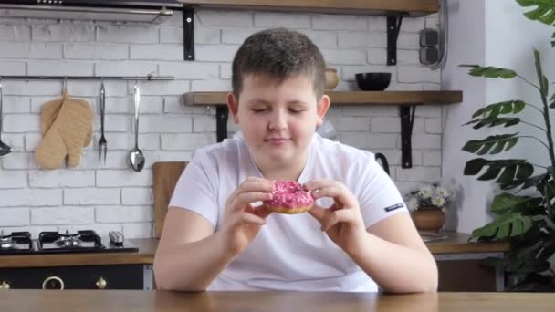 The fat man happily eats a donut in the kitchen. Chubby teen eats a donut 4k - Metraje, vídeo