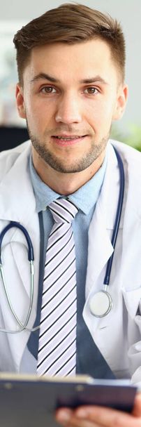 Ritratto di un bel dottore sorridente che lavora in clinica. Medico cardiologo terapeuta scegliere il giusto metodo di trattamento e diagnosi medica seduto a tavola con scheda paziente - Foto, immagini