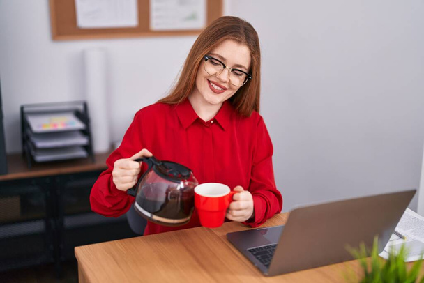 Jonge roodharige vrouw zakenman gieten koffie op beker op kantoor - Foto, afbeelding