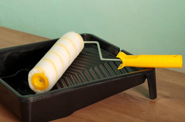 Paint roller with a yellow handle and a black plastic paint tray on wooden board. Building, renovation and construction work. Tools for painting walls: roller, tray near a green wall. Interior update - Foto, afbeelding