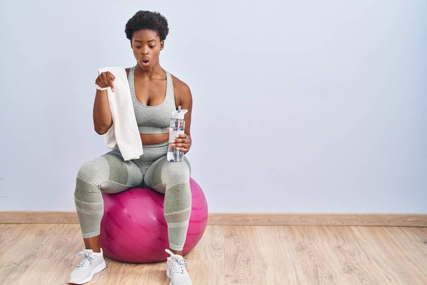 African american woman wearing sportswear sitting on pilates ball pointing down with fingers showing advertisement, surprised face and open mouth  - Foto, Bild